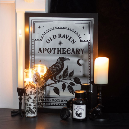 Decorative sign for 'Old Raven Apothecary' with candles and bottles on a dark surface.