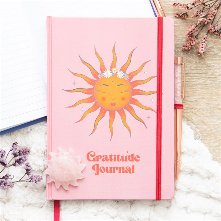 Pink gratitude journal with a sun illustration on a white surface with flowers.