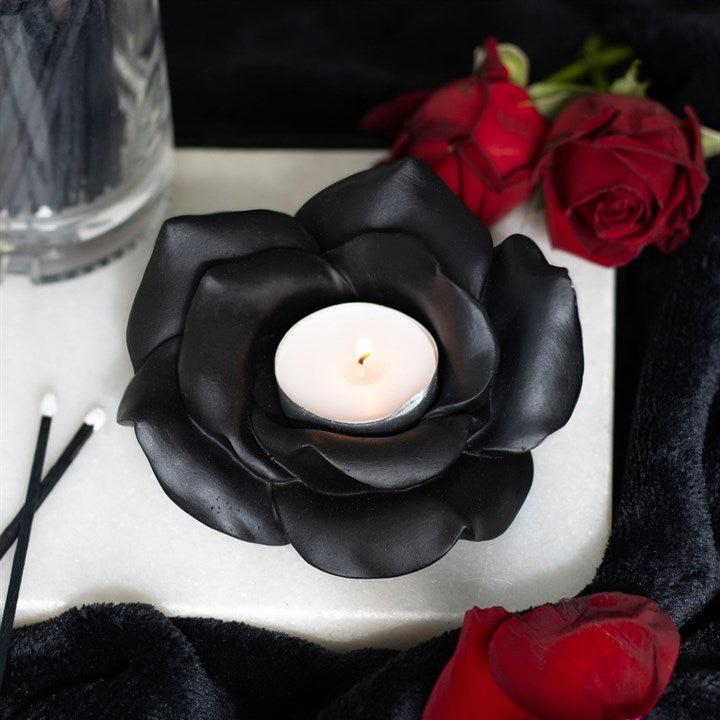 Black rose-shaped candle holder with a white candle, surrounded by red roses on a dark surface.