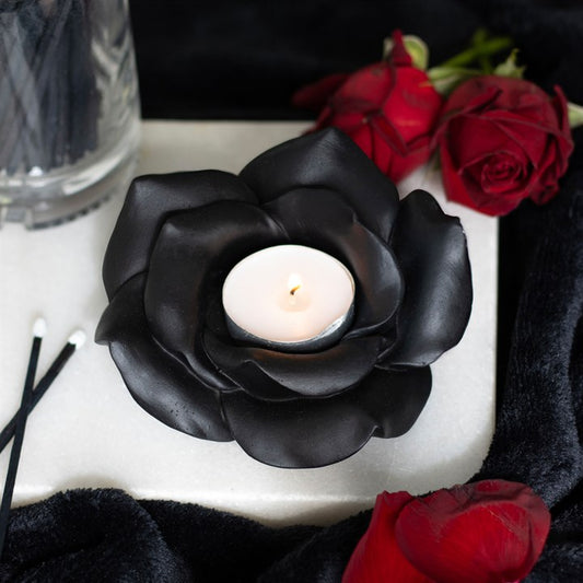 Black rose-shaped candle holder with a white candle, surrounded by red roses on a dark surface.