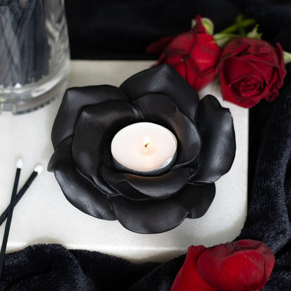 Black rose-shaped candle holder with a white candle, surrounded by red roses on a dark surface.