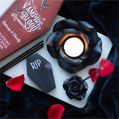 Black rose-shaped candle holder with a lit candle, 'Vampire Blood' book, and 'RIP' card on a dark surface.