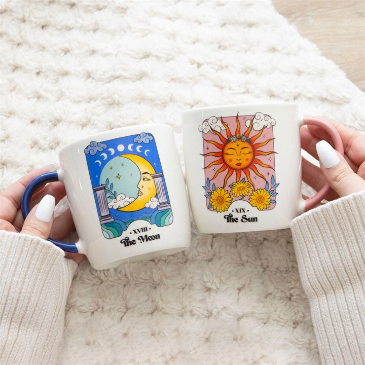 Two tarot-themed mugs held by hands on a textured surface