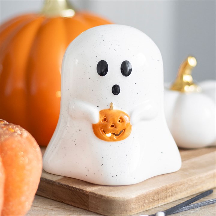 Ghost-shaped candle holder with pumpkin face design on a wooden board with pumpkins in the background