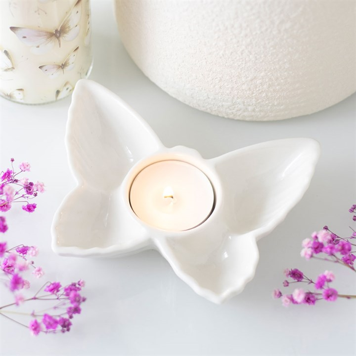 White ceramic butterfly-shaped candle holder with a lit tealight candle on a light background with pink flowers.
