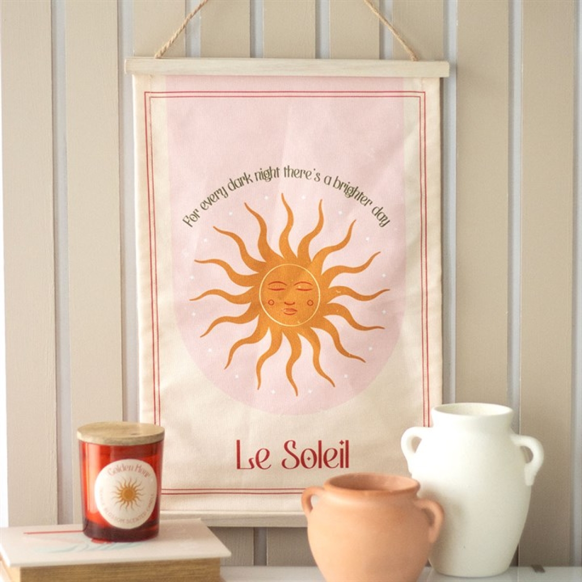 Decorative wall hanging with sun illustration and text, placed on a shelf with a candle and vase.
