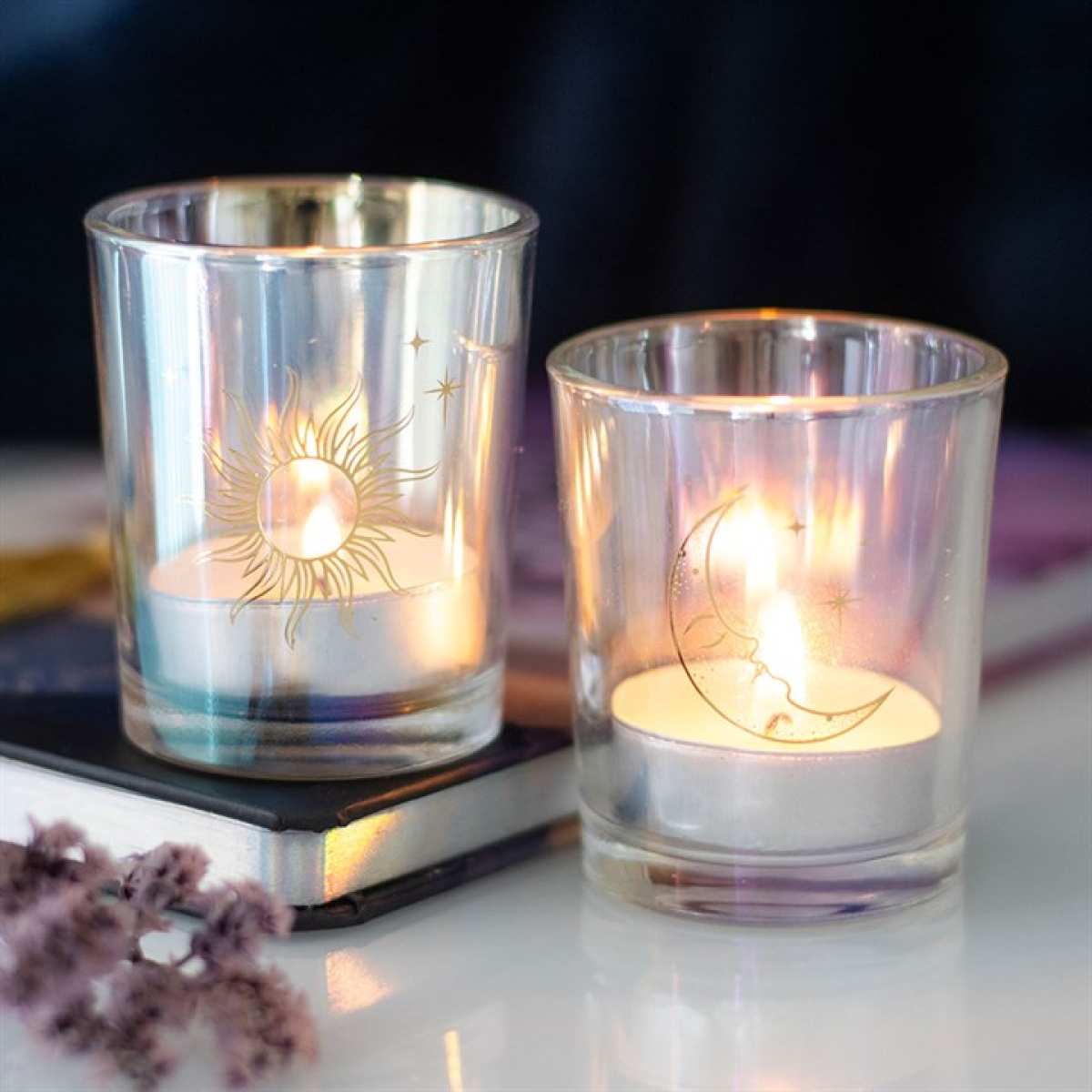 Two glass candle holders with engraved designs of the sun and moon, containing lit candles on a reflective surface.