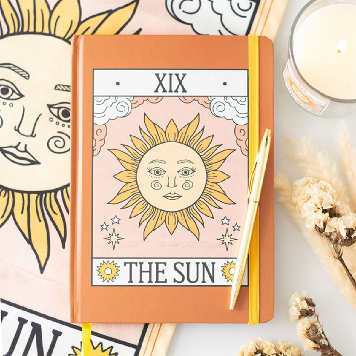 Notebook with 'The Sun' tarot card design on a table with candles and flowers.