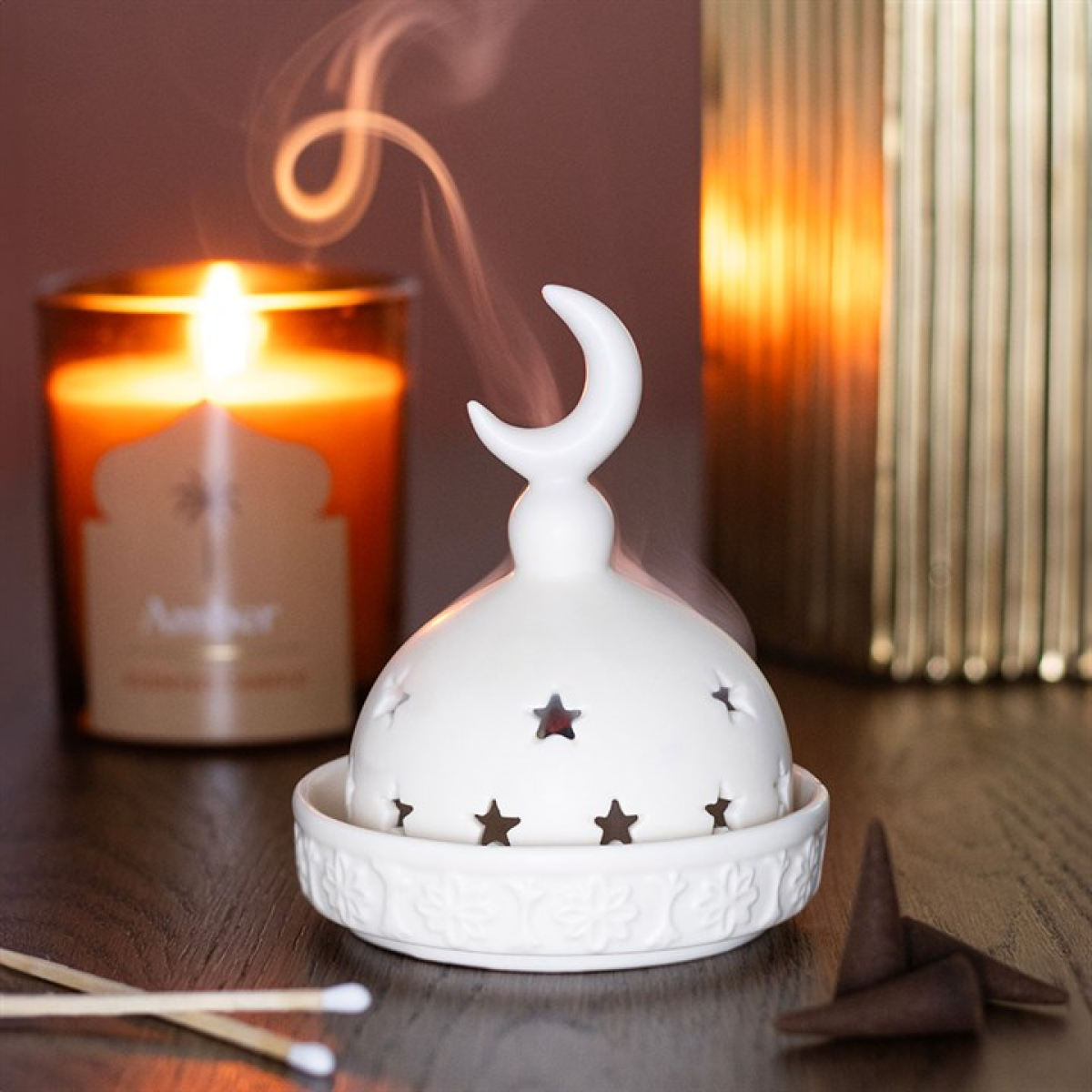 White ceramic incense burner with star designs on a wooden surface, accompanied by a lit candle and a book.